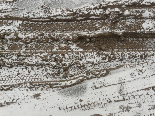 Dirt road texture in dark brown tone with tire marks and thin layer of snow across surface.